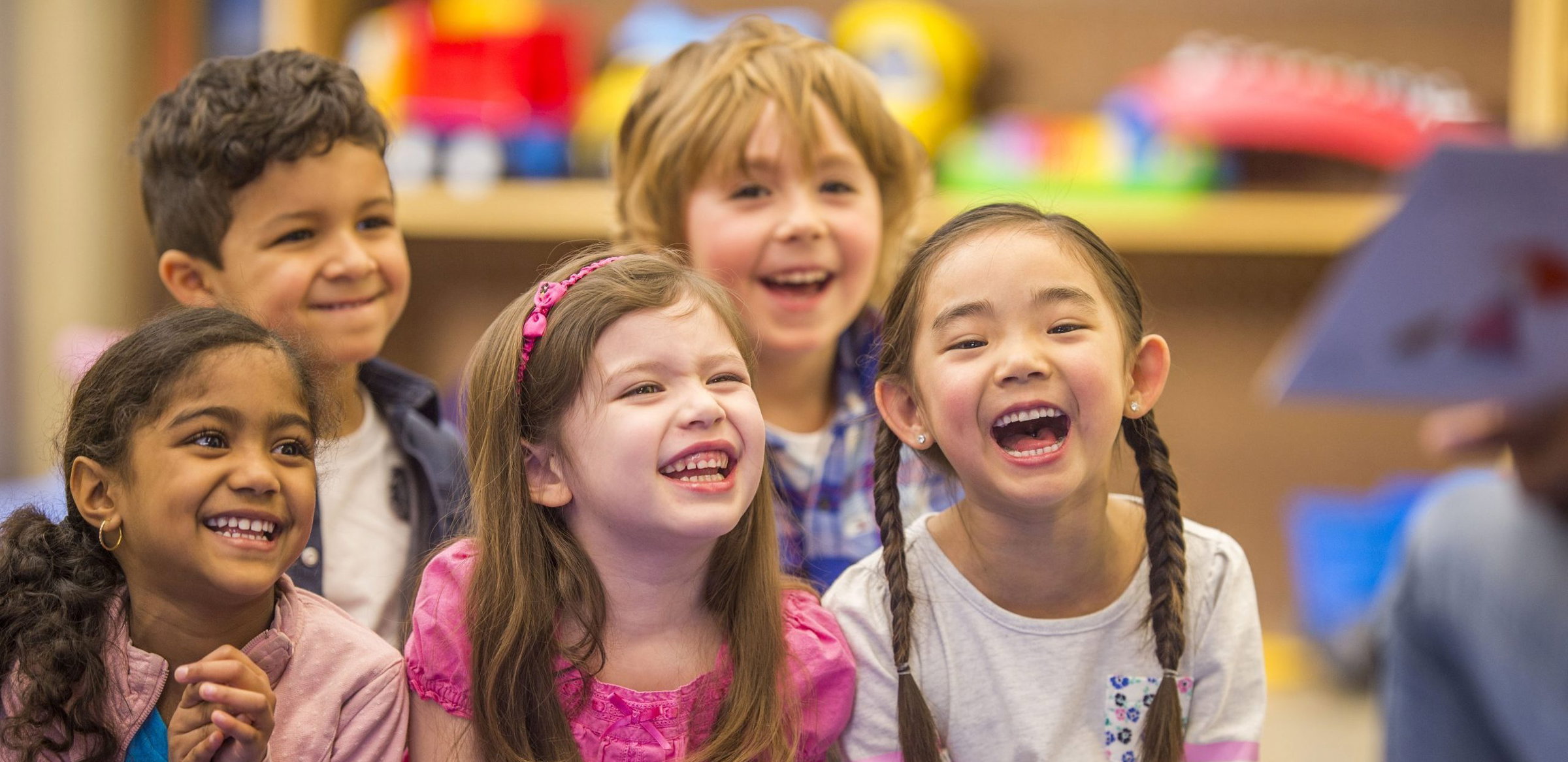 A multi-ethnic group of elementary age children are laughing while their teacher reads them a book during story time.
